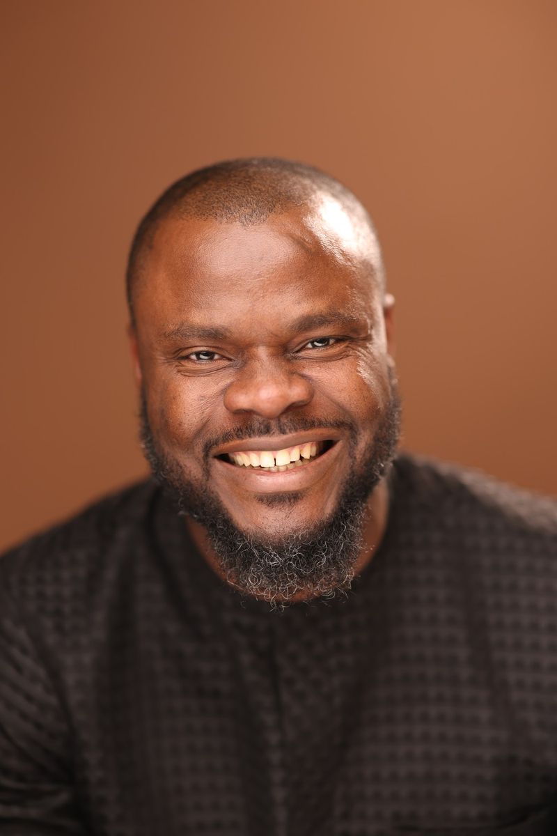 Elvis C. Umez smiling, in a studio portrait against a warm brown background