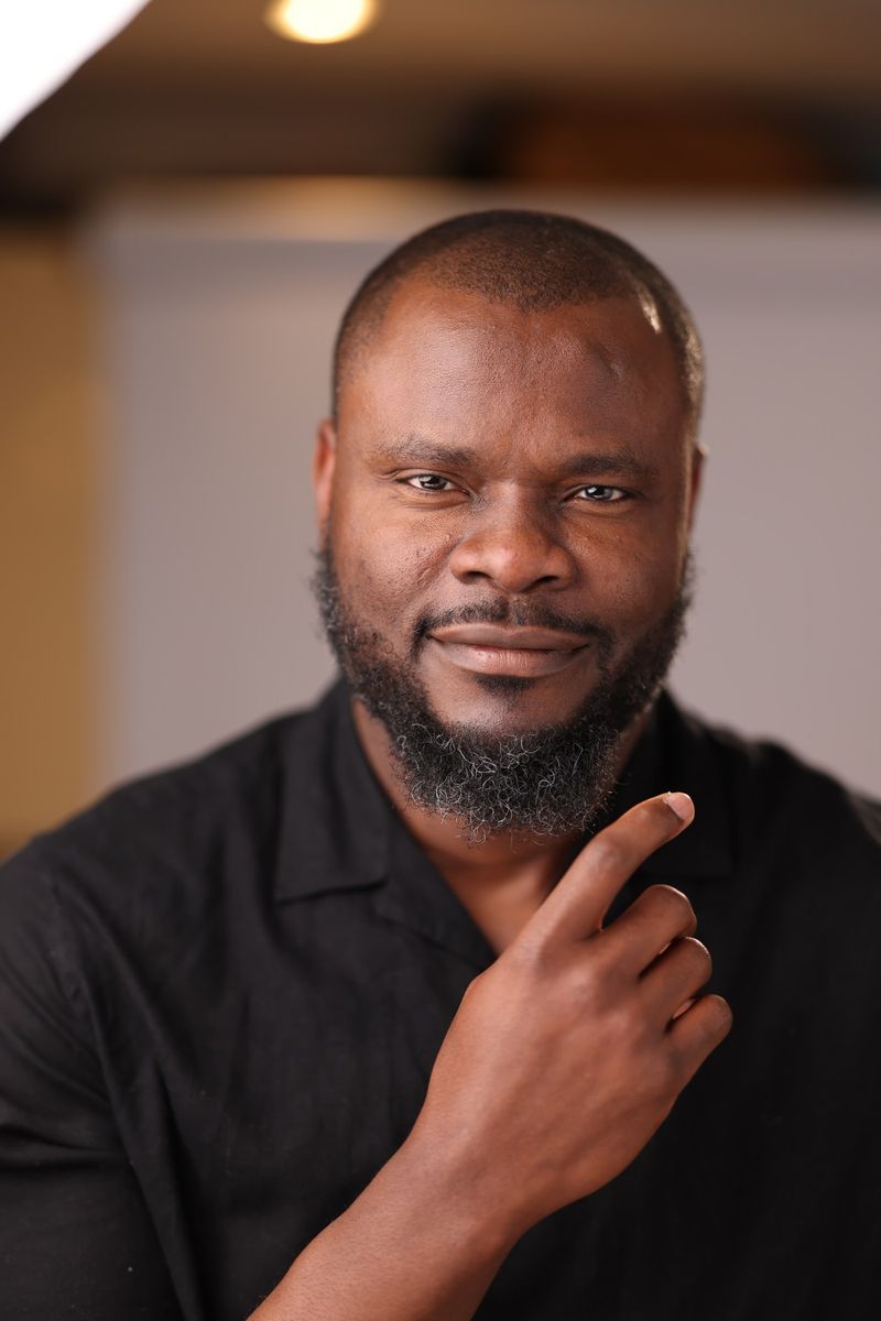 Elvis C. Umez in a black shirt, one hand raised to his chin, looking at the camera